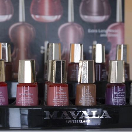 Eine Ausstellung von Mavala Nagellacken in verschiedenen Farben, einschließlich Hortensi, Freesia, Cairo und Jelly Grape. Die Flaschen befinden sich auf einem schwarzen Halter mit dem Markennamen Mavala und dem Herkunftsland Schweiz.