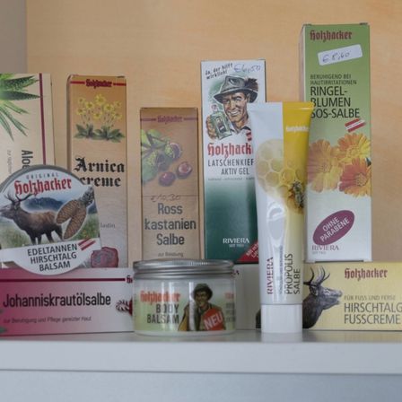 Verschiedene Hautpflegeprodukte wie Cremes, Gele und Balsame sind auf einem Regal angeordnet. Produkte umfassen Arnika-Creme, Ross Kastanien Salbe und Johanniskrautölsalbe. Andere Artikel sind auf Deutsch beschriftet.