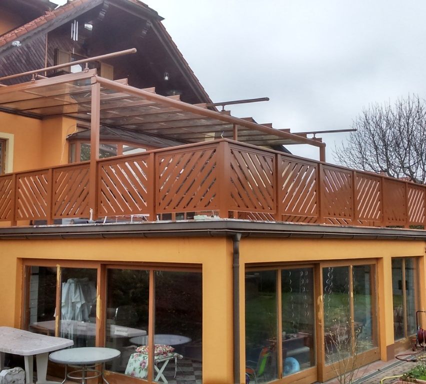 Ein gelbes Haus mit einem braunen Balkon und einer Metallüberdachung. Im Inneren gibt es Tische und Stühle. Draußen ein Baum und ein grüner Rasen.