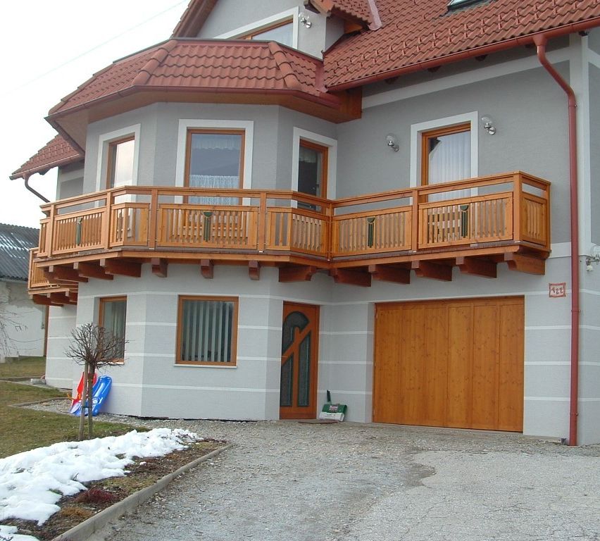 Ein zweistöckiges Haus mit rotem Dach und Holzbalkon, einer hölzernen Garagentür und einem kleinen Baum davor. Schnee liegt auf dem Boden.