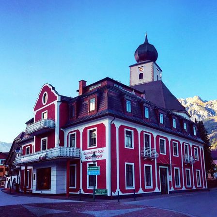 Ein rot-weißes Gebäude mit einem Turm, benannt Gesundheit Hotel Sparger. Es hat mehrere Fenster und Balkone. Berge sind in der Ferne.
