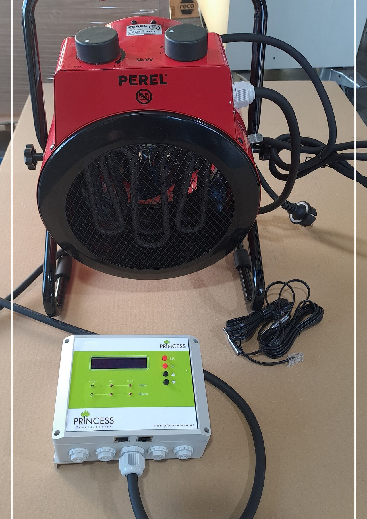 Ein roter und schwarzer Perel-Heizlüfter steht auf einer braunen Oberfläche. Er hat ein Gittergitter vorne und mehrere angeschlossene Kabel. Darunter befindet sich ein elektronisches Gerät der Marke Princess mit digitalem Display und Tasten.