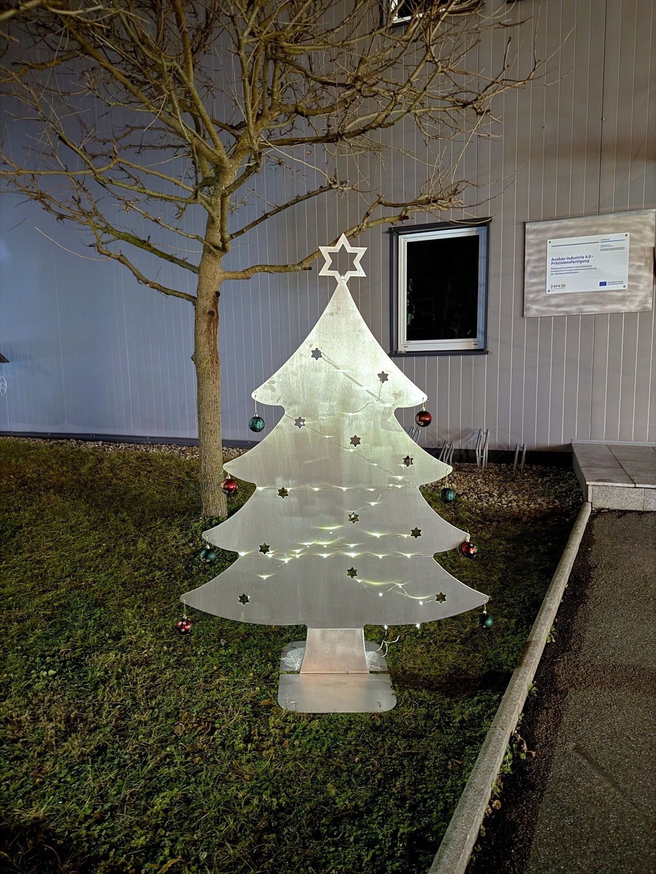 Ein dekorativer Metall-Weihnachtsbaum mit Lichtern und Ornamenten steht auf dem Rasen vor einem Gebäude mit Fenster und Schild.