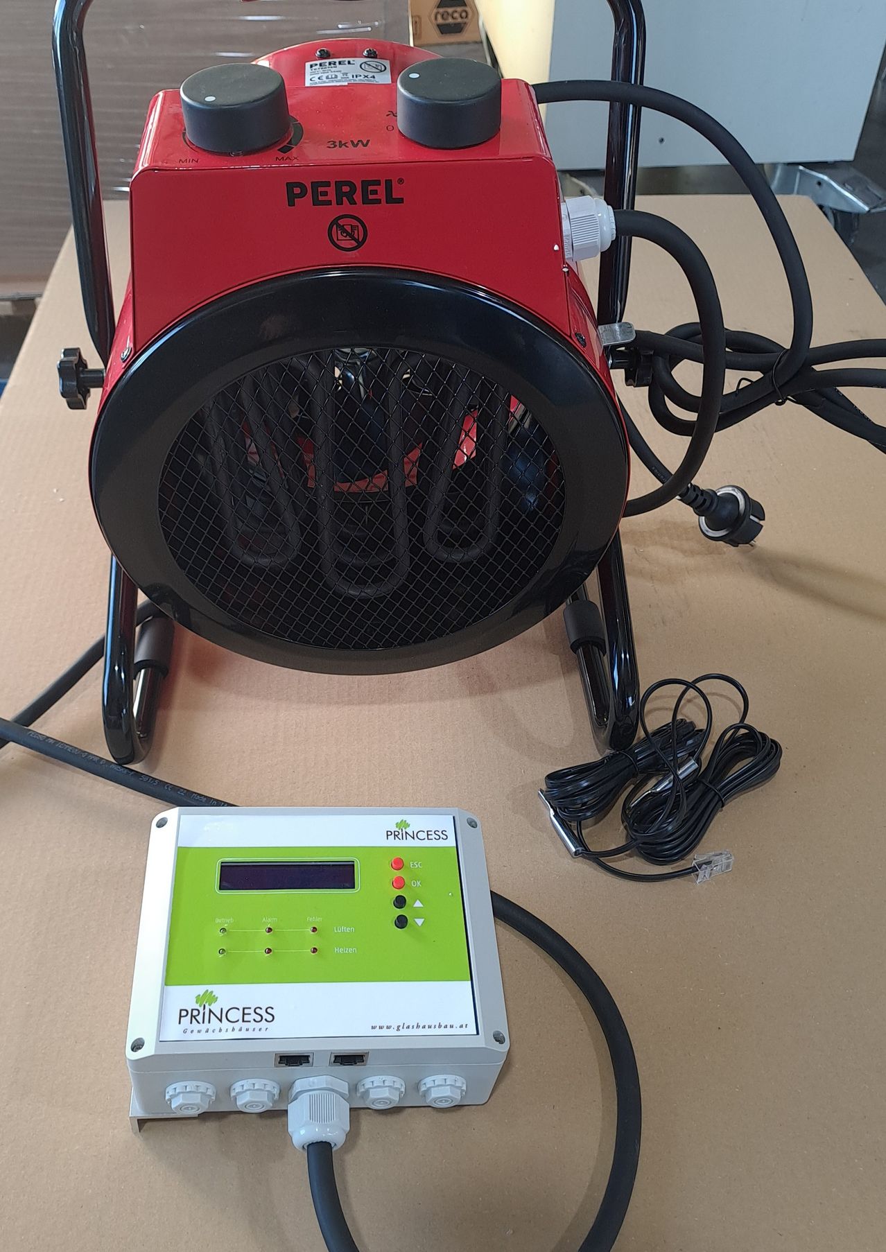 Ein roter elektrischer Perel-Heizlüfter mit digitalem Bedienfeld wird auf einer Pappunterlage ausgestellt. Der Heizlüfter hat ein Gittergitter und Steuerknöpfe.