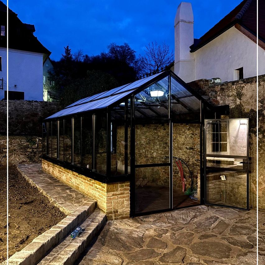 A modern greenhouse with a glass roof and brick base is illuminated at night, adjacent to a stone wall and a white building.