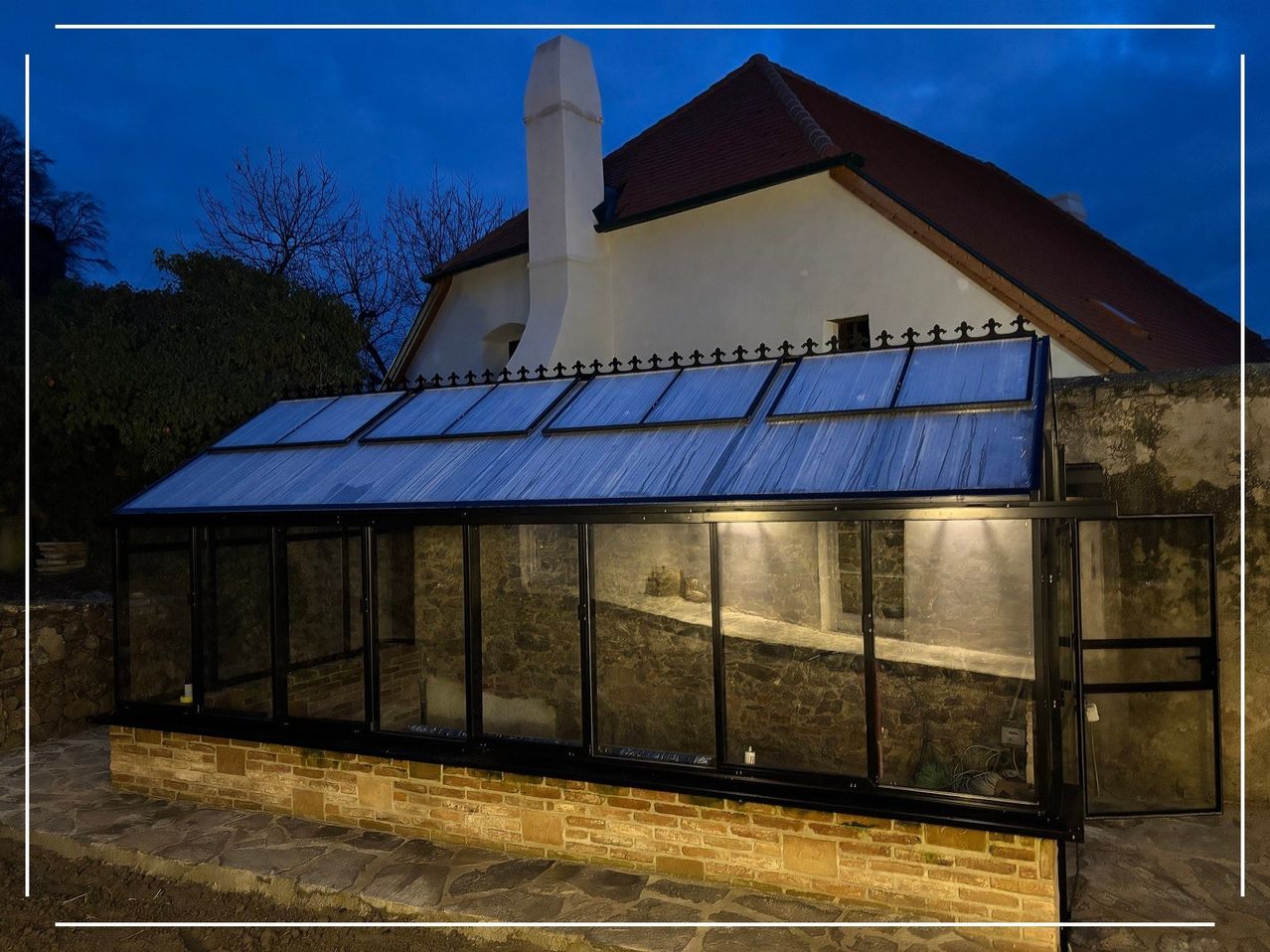 A greenhouse with a blue roof and glass walls is attached to a brick building under a clear night sky.