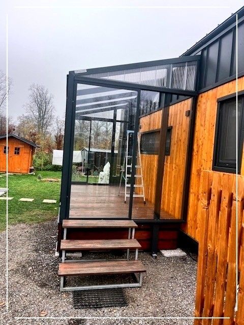 Ein modernes Holzhaus mit einer Glasüberdachung, Stufen und einer Leiter. Im Hintergrund befinden sich ein kleines Haus und ein Baum.