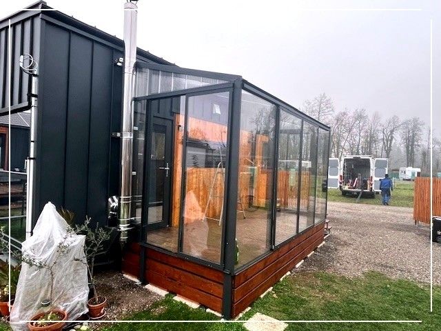 Ein schwarzes Gebäude mit einer Glasverlängerung, einem überdachten Gang und einer Leiter. Bäume und ein Van im Hintergrund.