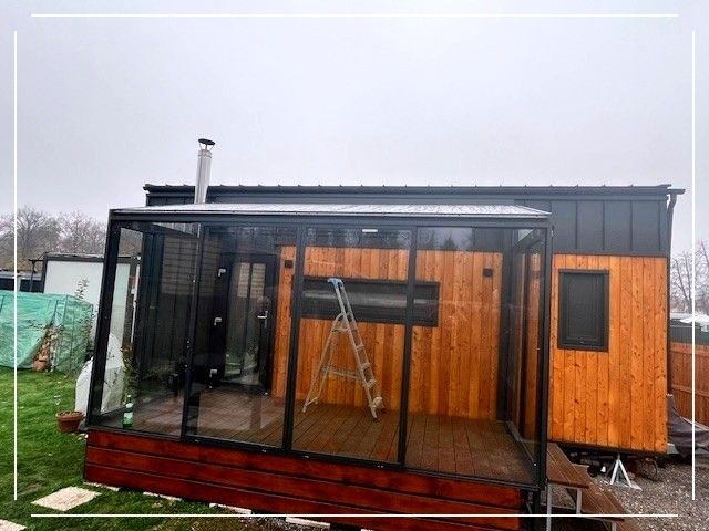Ein modernes Tiny House mit hölzerne Außenwand und einer verglasten Veranda. Es hat ein schräges Dach und einen Schornstein. Eine Leiter steht in der Veranda.