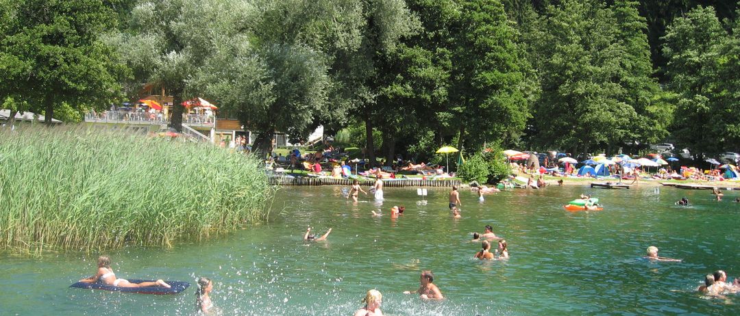 Menschen schwimmen in einem von Grün umgebenen See, einige sonnen sich am Ufer und andere entspannen unter Sonnenschirmen. Bäume und Berge bilden den Hintergrund.