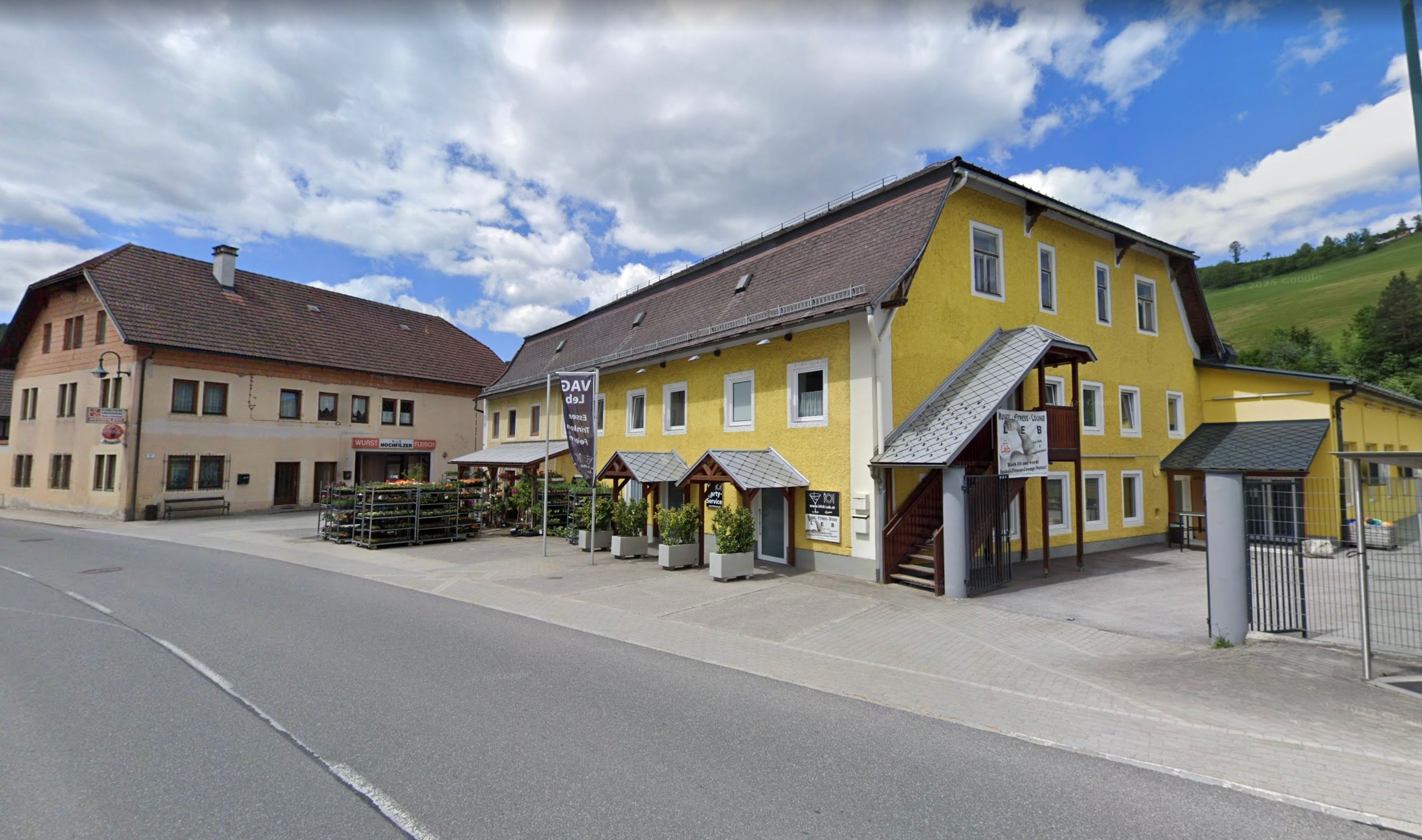 Ein leuchtend gelbes Gebäude mit braunem Dach steht am Straßenrand. Vorne stehen Pflanzen und ein Balkon über dem Eingang.