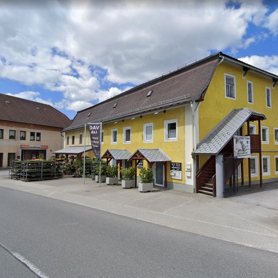 Ein gelbes Gebäude mit einem braunen Dach steht vor einer Straße. Ein Schild an der Seite steht für 'AV'. Vor dem Gebäude stehen Pflanzen. Eine Treppe führt zu einem Balkon.