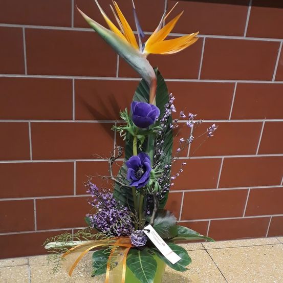 Eine Blumenarrangement mit leuchtend lila und gelben Blumen ist in einem grünen Gefäß vor einer Ziegelwand ausgestellt.