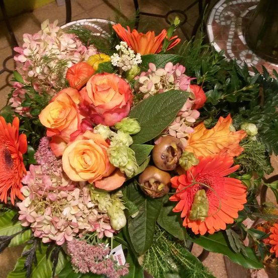 Eine lebendige Blumenzusammenstellung mit orangen und rosa Blumen, einschließlich Rosen, Hortensien und Ringelblumen, mit grünen Blättern und dekorativen Kürbissen.