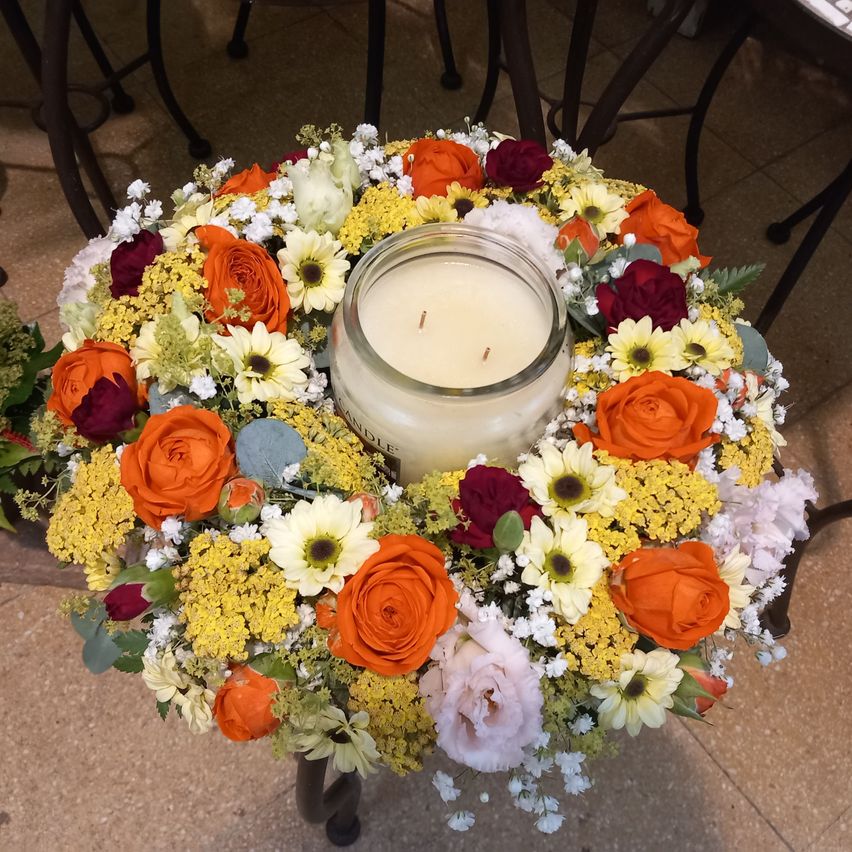 Ein Kranz aus orangefarbenen, gelben und roten Blumen umgibt eine weiße Kerze auf einer gefliesten Oberfläche.