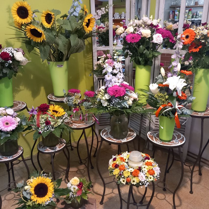 Ein Raum voller verschiedener Blumenarrangements, darunter Sonnenblumen, Gänseblümchen und gemischte Blumen, die auf grünen Vasen auf kleinen Tischen vor einer gelben Wand ausgestellt sind.