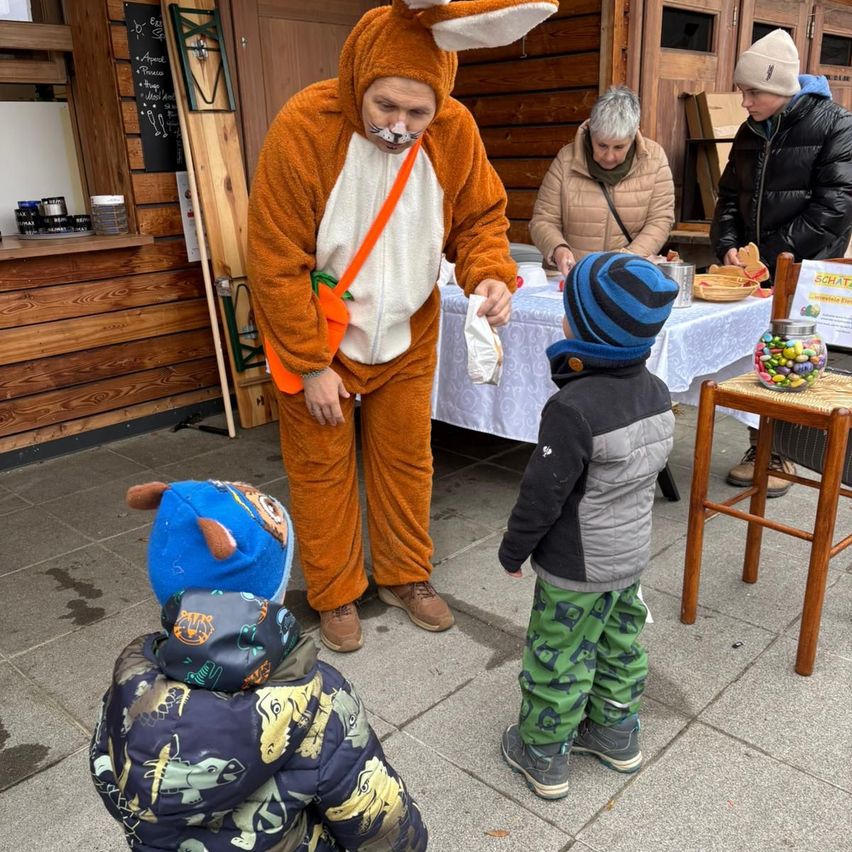 A man in a fox costume interacts with two children. One child looks at the man while the other examines a table with candies and a jar. In the background, two adults observe.