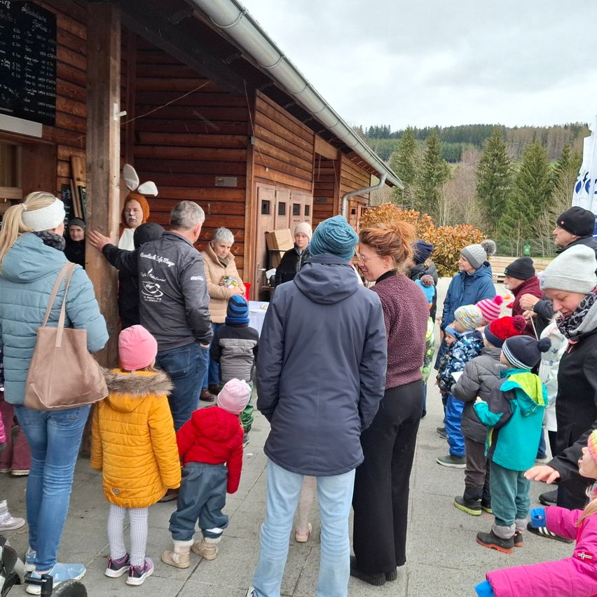 A group of people, including children, stand in line outside a wooden building. Some are wearing hats and coats. A woman in a pink hat looks at a child in a yellow jacket.
