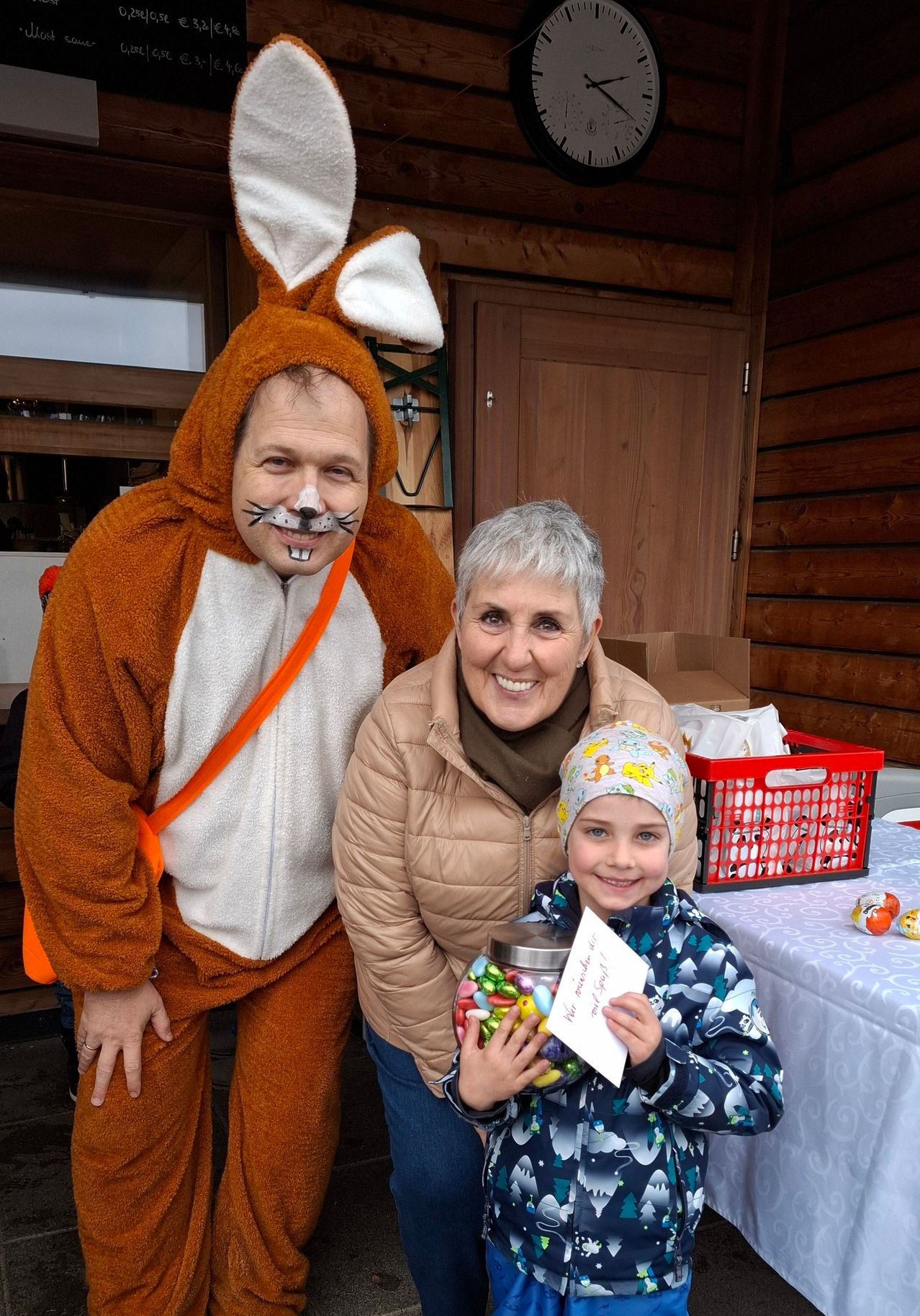 A family poses for a photo with a man in a rabbit costume, a smiling woman holding a jar of candy, and a child in a blue jacket with a tag in front of a wooden wall.