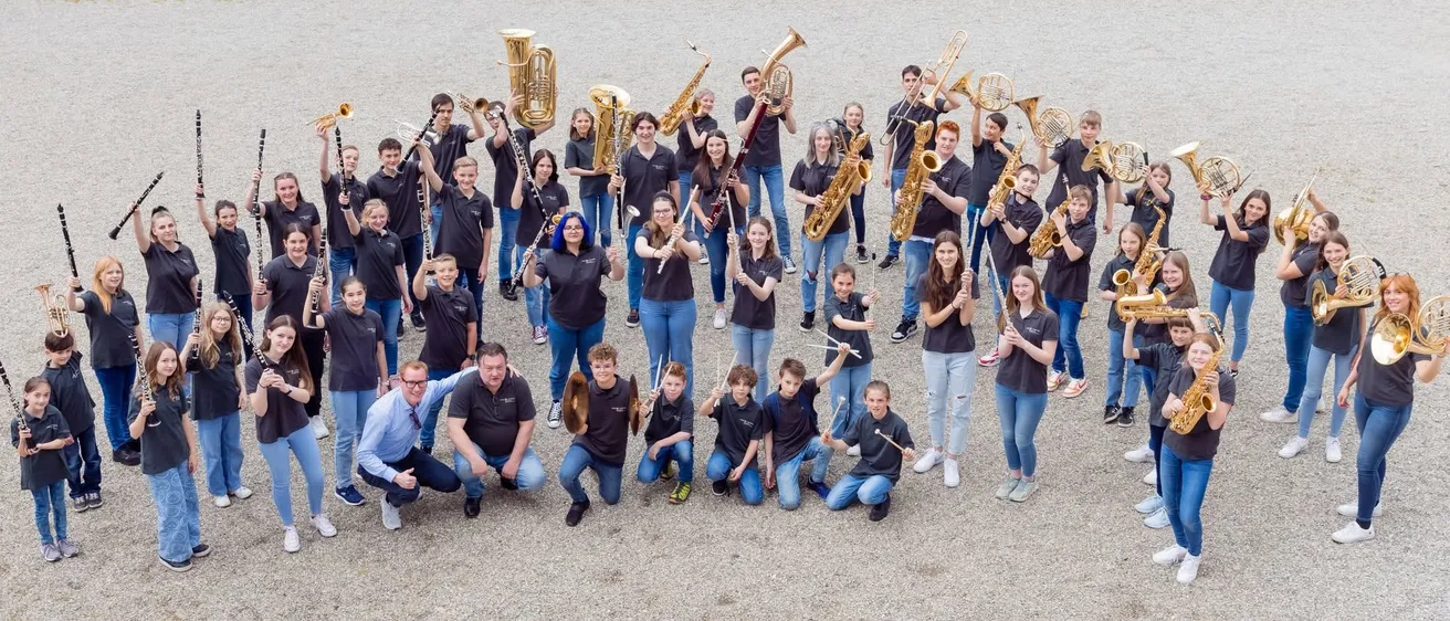 Eine Gruppe junger Musiker in schwarzen T-Shirts posiert mit verschiedenen Blech- und Holzblasinstrumenten für ein Foto. Sie stehen und knien auf einer Kiesfläche.