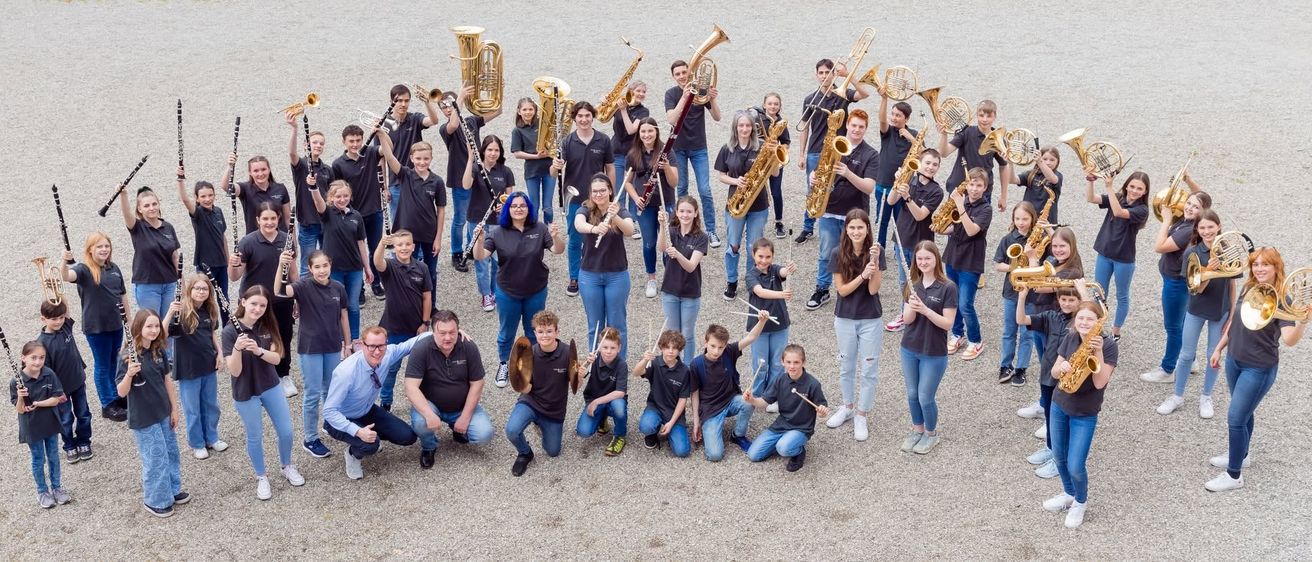 Eine Gruppe junger Musiker in schwarzen T-Shirts posiert mit verschiedenen Blech- und Holzblasinstrumenten für ein Foto. Sie stehen und knien auf einer Kiesfläche.