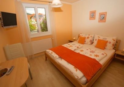 Ein gemütliches Schlafzimmer mit einem Holzbett, weißen Laken, orangefarbenen und weißen Kissen, einer Fernbedienung auf einem Holztisch, einem an der Wand montierten Fernseher und einem weißen Stuhl.