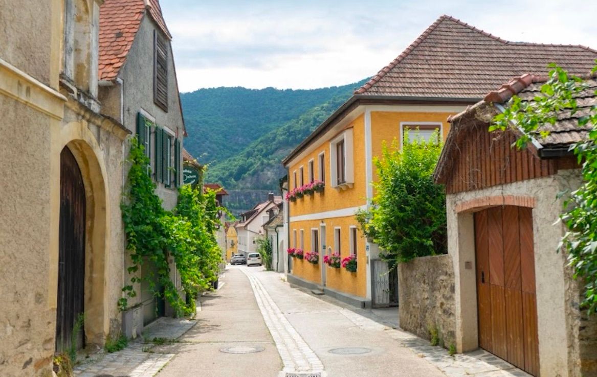 Eine schmale Straße gesäumt von gelben Häusern, eines mit rosa Blumen, unter einem bewölkten Himmel, mit einem Berg im Hintergrund.