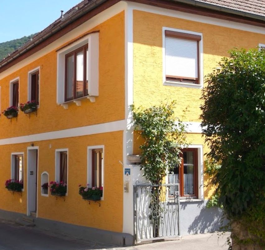 Ein gelbes Haus mit weißen Zierleisten hat mehrere Fenster, Blumenkästen und ein kleines Tor. Ein Baum wächst im Vordergrund.