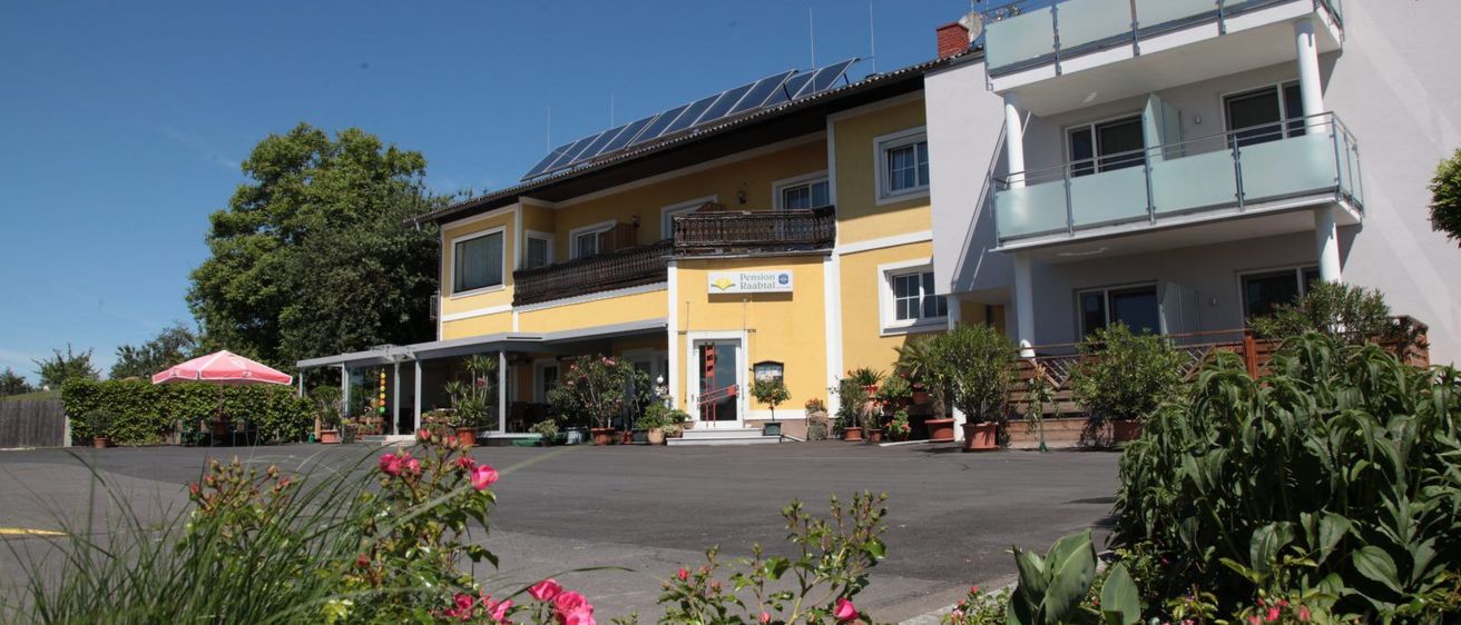 Ein gelbes Gebäude mit Solarpanelen auf dem Dach hat einen Balkon und mehrere Pflanzen. Es ist von Blumen und Grün umgeben, mit einem klaren blauen Himmel im Hintergrund.