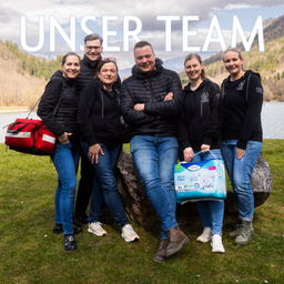Eine Gruppe lächelnder Personen, die schwarze Jacken und Jeans tragen, stehen und sitzen auf einem großen Felsen in einer malerischen Außenumgebung. Der Text 'UNSER TEAM' ist prominent über ihnen angezeigt.