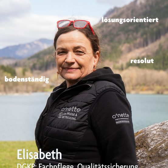 Eine Frau namens Elisabeth steht im Freien und lächelt, mit einem See und Bergen im Hintergrund. Sie trägt eine schwarze Jacke mit weißem Text und roten Brillen auf dem Kopf.
