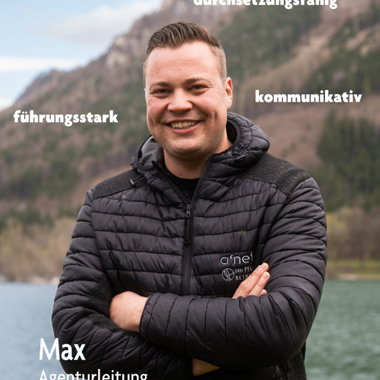 Ein Mann in einer schwarzen Jacke mit einem Logo auf der linken Brust posiert lächelnd für ein Foto mit verschränkten Armen. Im Hintergrund befinden sich ein See und Berge.