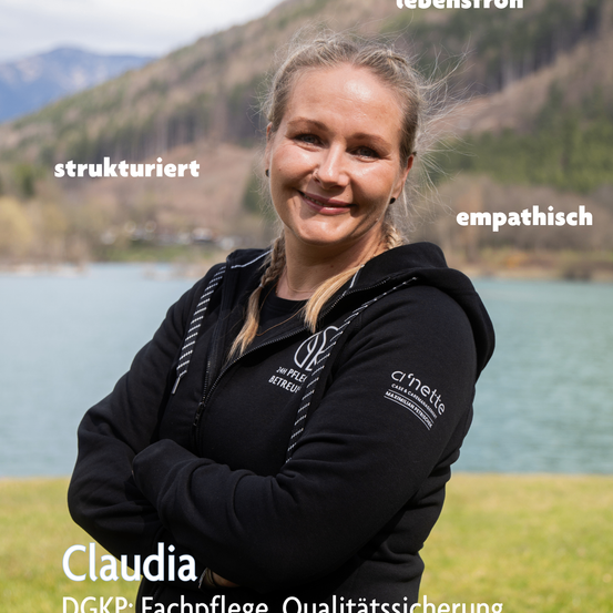 Eine Frau namens Claudia steht vor einem See mit Bergen im Hintergrund. Sie trägt eine schwarze Kapuzenjacke mit dem Logo einer Firma namens Clinette. Sie lächelt und scheint für ein Foto zu posieren.