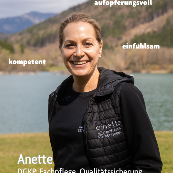 Eine lächelnde Frau namens Anette, die eine schwarze Weste mit dem Logo '24h Pflege & Betreuung' trägt, steht in der Nähe eines Sees mit Bergen im Hintergrund.