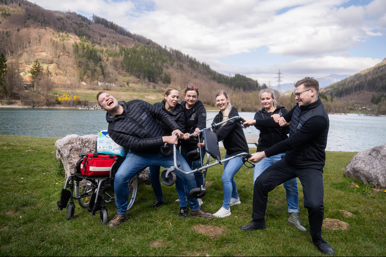 Eine Gruppe von Menschen in schwarzen Jacken und Jeans hat Spaß mit einem Rollstuhl auf einem Grasfeld in der Nähe eines Sees mit Bergen im Hintergrund.