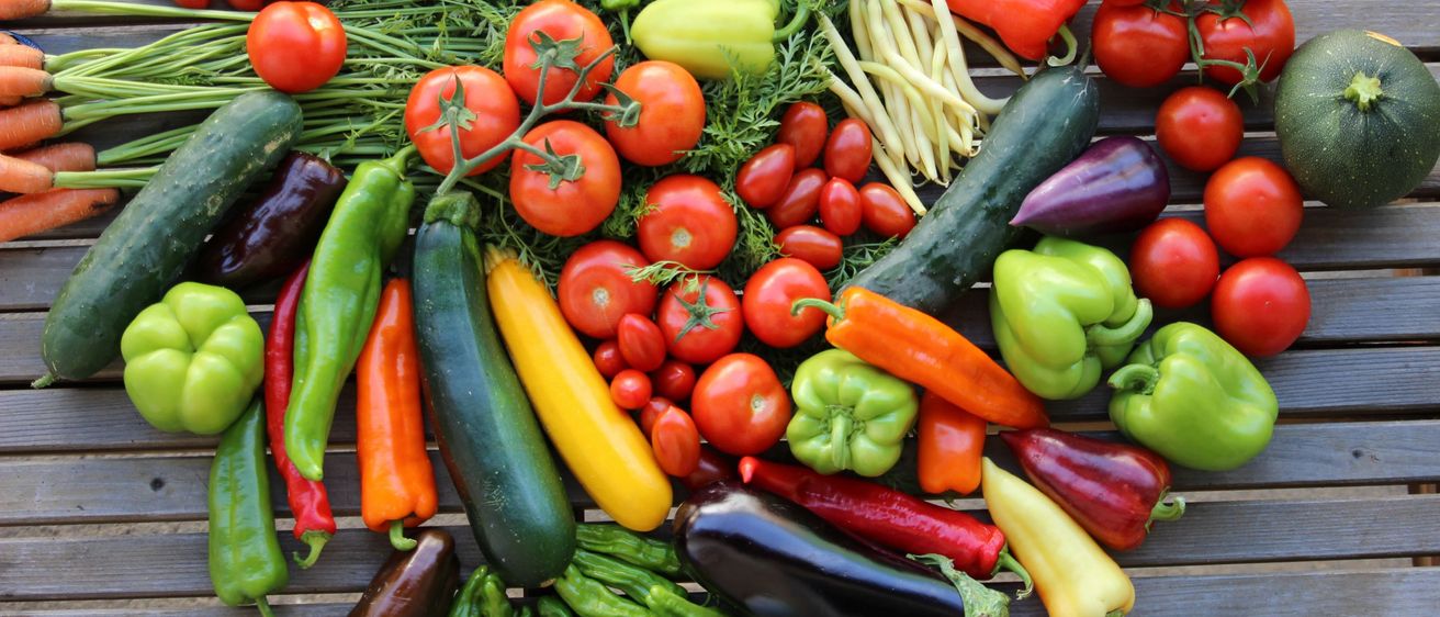 Eine Vielzahl von Gemüsesorten wie Tomaten, Paprika, Auberginen, Gurken und Bohnen sind auf einer Holzoberfläche angeordnet.