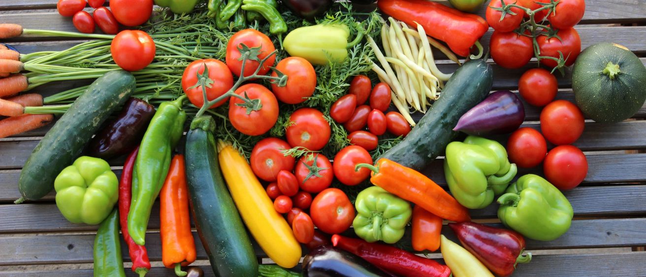Eine Vielzahl von Gemüsesorten wie Tomaten, Paprika, Auberginen, Gurken und Bohnen sind auf einer Holzoberfläche angeordnet.