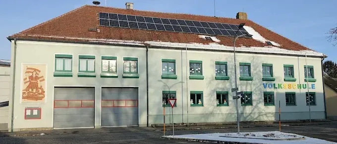 Das Volksschule-Gebäude hat ein braunes Dach mit Solarpaneelen. Es hat eine Wandmalerei, Doppelgaragentore und viele Fenster.