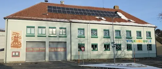 Das Volksschule-Gebäude hat ein braunes Dach mit Solarpaneelen. Es hat eine Wandmalerei, Doppelgaragentore und viele Fenster.