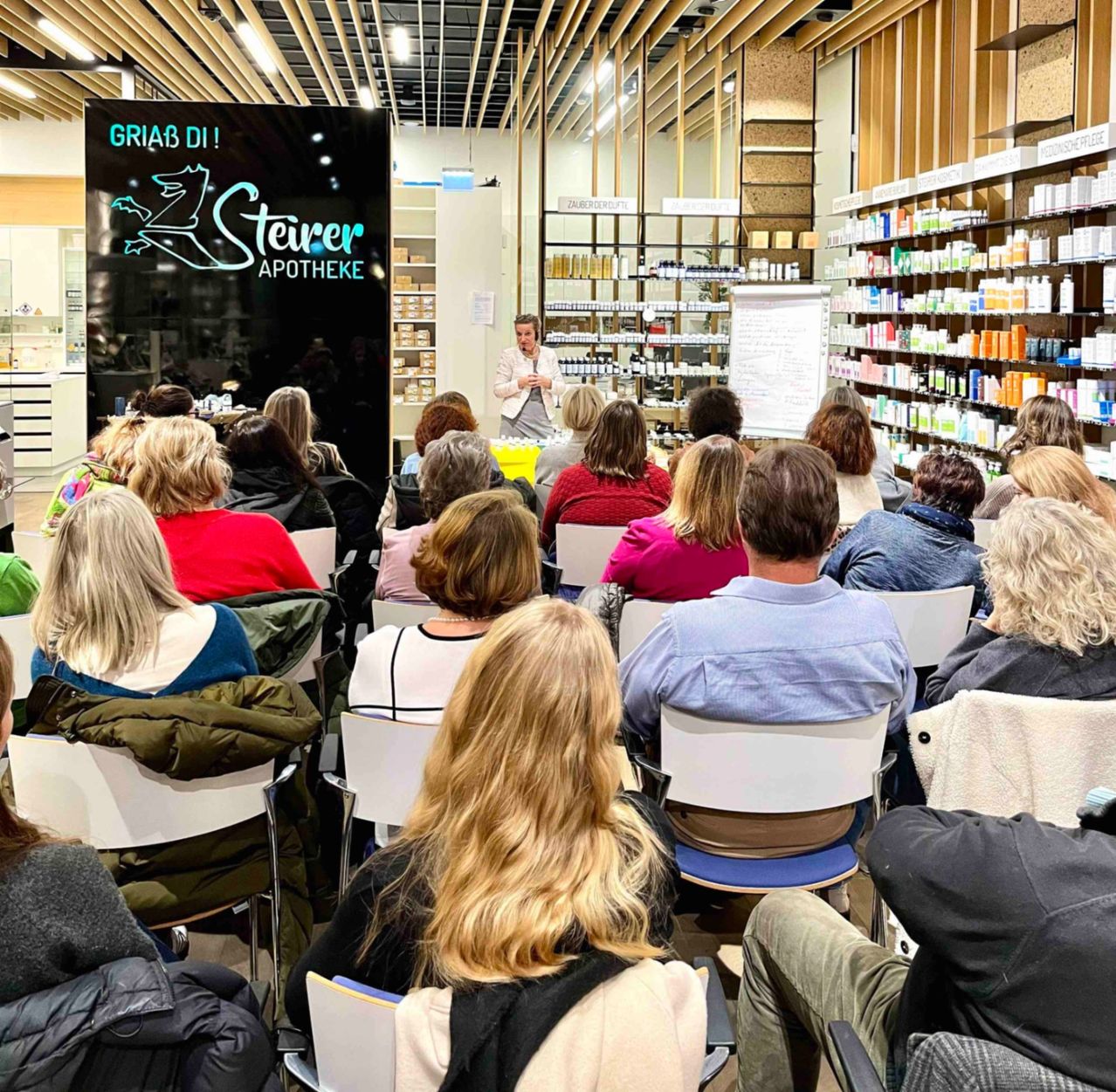Eine Veranstaltung in einer Apotheke, bei der eine Frau vor einem Publikum spricht. Das Publikum sitzt in Stühlen und schaut ihr zu. Hinter ihr befindet sich ein Schild mit 'Steiver Apotheke' und Regale gefüllt mit Produkten.