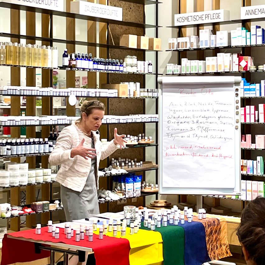 Eine Frau in einer weißen Blazerjacke und einer grauen Hose hält eine Präsentation in einem Raum mit Regalen voller Produkte. Der Tisch vor ihr hat verschiedene kleine Flaschen. Dahinter zeigt eine Tafel handgeschriebenen Text. Regale auf beiden Seiten sind mit zahlreichen Flaschen und Kartons bestückt.