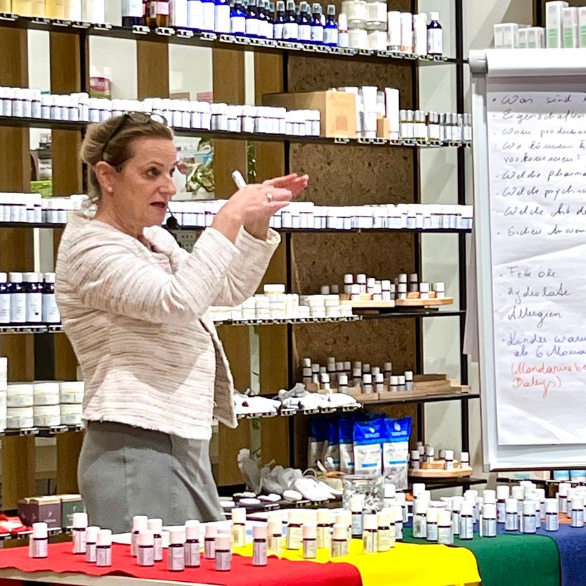 Eine Frau hält einen Vortrag über ätherische Öle und steht vor Regalen mit verschiedenen Flaschen und Behältern. Sie hält einen Marker und zeigt auf eine Tafel.