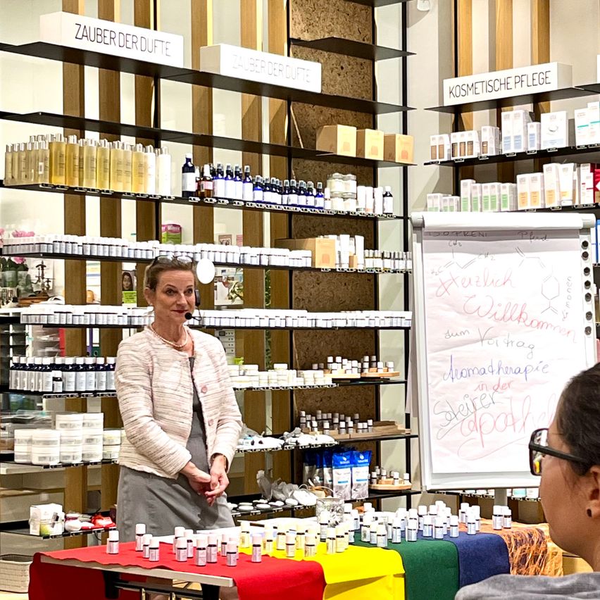 Eine Frau steht vor einem Geschäft und präsentiert ätherische Öle. Hinter ihr sind Regale mit Kosmetikprodukten gefüllt. Eine Tafel zeigt Informationen über Aromatherapie.