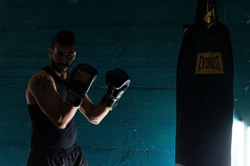 Ein Mann in einem schwarzen Tanktop und Handschuhen posiert vor einer blauen Wand mit einem schwarzen Boxsack, der mit Everlast beschriftet ist.