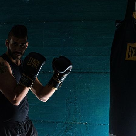 Ein Mann in einem schwarzen Tanktop und Handschuhen posiert vor einer blauen Wand mit einem schwarzen Boxsack, der mit Everlast beschriftet ist.