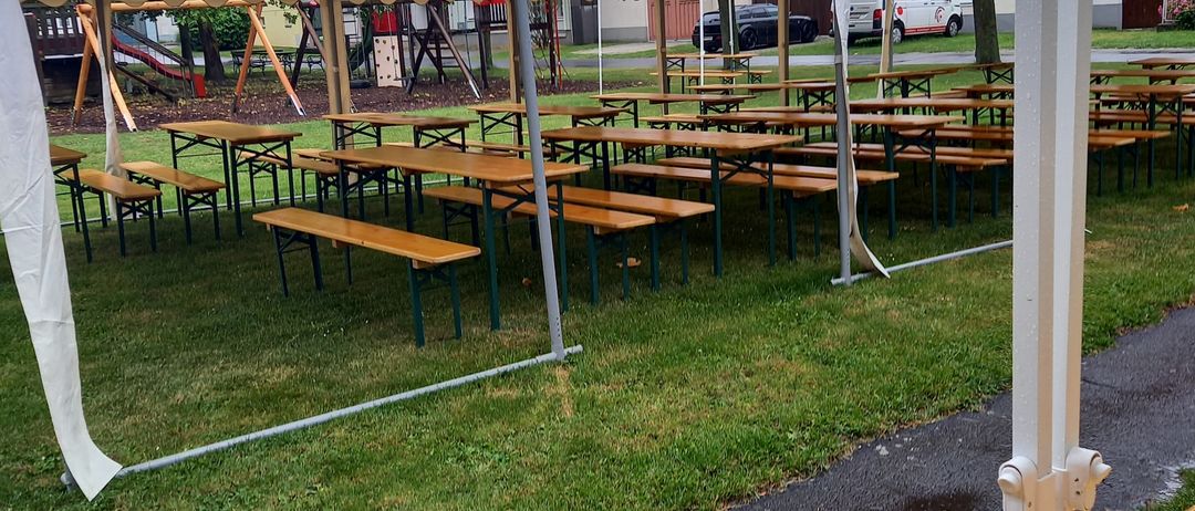 Ein Freisitzbereich mit Holztischen und Bänken, bedeckt von einem weißen Zelt. Der Bereich ist von Gras und einem Betonweg umgeben. Bäume und ein Spielplatz sind im Hintergrund.