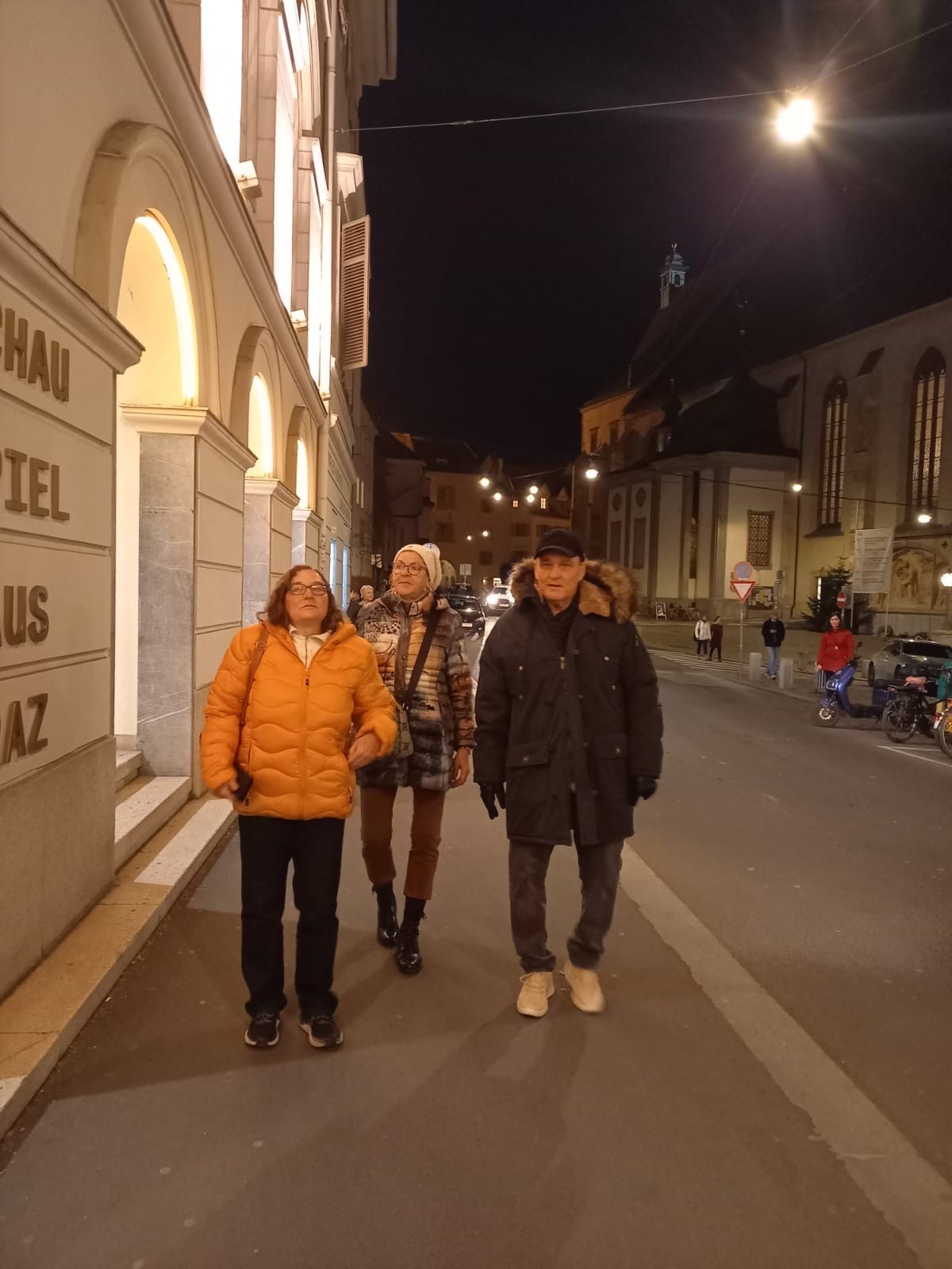 Drei Personen gehen nachts auf einer Straße. Ein Gebäude mit dem Wort 'Schauspielhaus' darauf. Eine Kirche befindet sich auf der rechten Seite. Es gibt geparkte Autos und Menschen, die auf der Straße gehen.