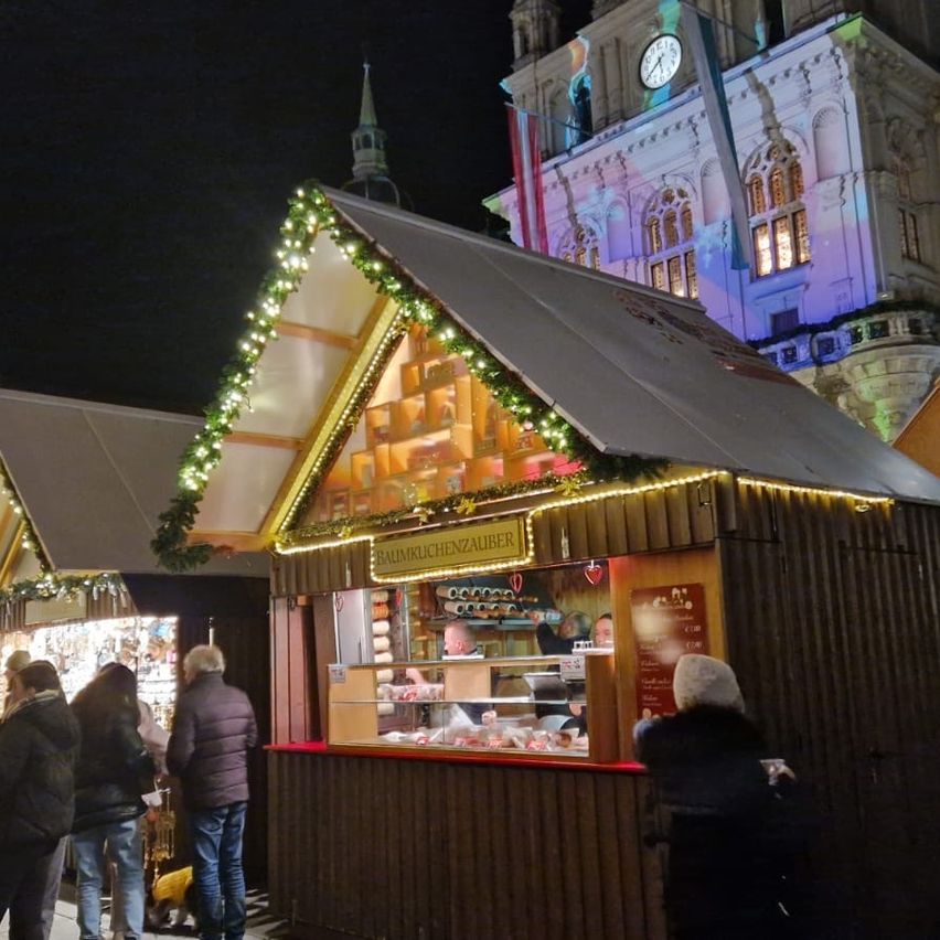 Bei Nacht ist ein festlich geschmückter Weihnachtsmarktstand voller Aktivität. Menschen versammeln sich und genießen die festliche Atmosphäre.