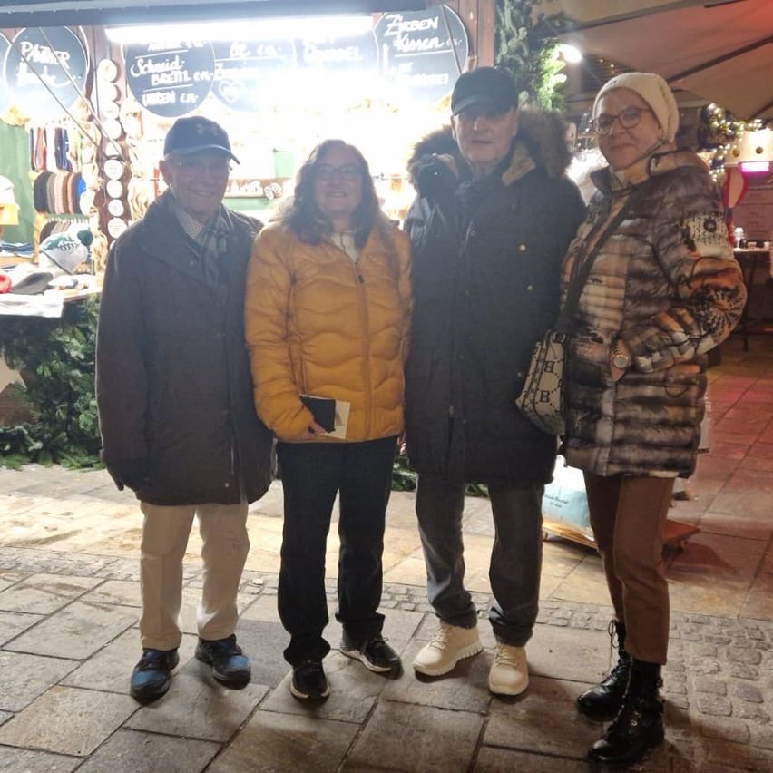 Vier Menschen stehen nachts draußen und lächeln für ein Foto. Sie sind alle warm gekleidet. Hinter ihnen befindet sich ein Marktstand mit verschiedenen zum Verkauf stehenden Artikeln.