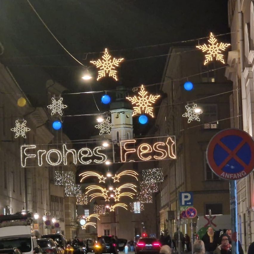 Eine Stadtstraße bei Nacht mit festlichen Dekorationen. Schneeflocken und blaue Lichter schmücken die Straße. Ein Schild sagt Frohes Fest. Fußgänger gehen auf dem Bürgersteig.